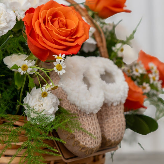 A pair of beige knit baby booties nestled in a wicker basket surrounded by orange roses and white daisies.