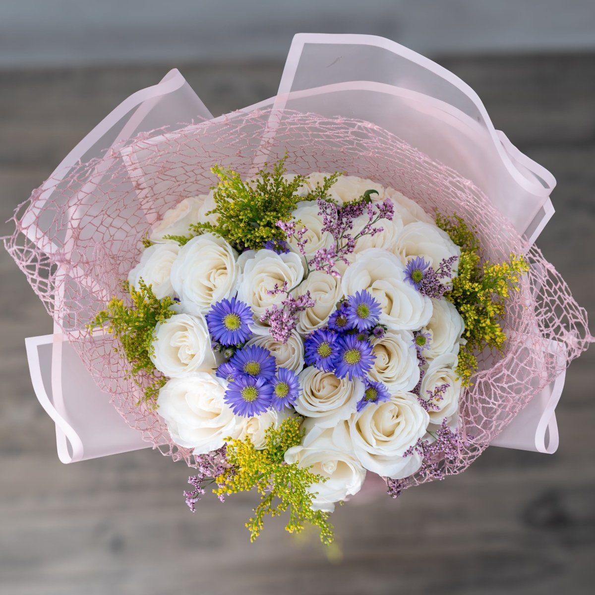 A bouquet of white roses and blue daisy-like flowers wrapped in sheer light pink tissue paper and netting, photographed from above on a blurred gray wooden surface.