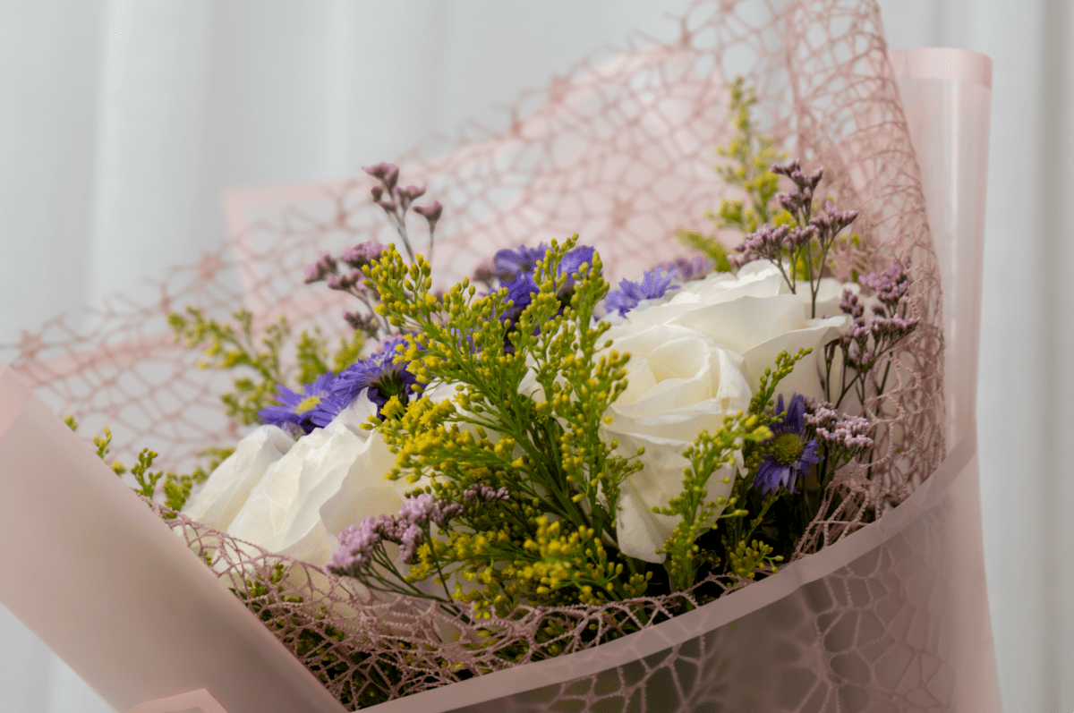 A close-up of a floral bouquet wrapped in pale pink mesh paper, featuring white roses, fillers, and greenery.