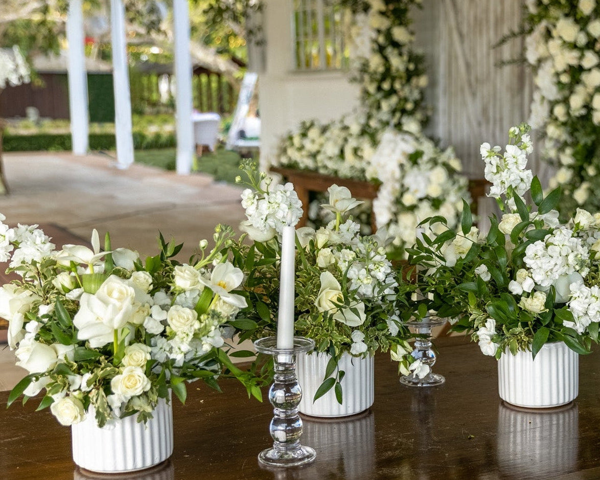 A long wooden dining table set with multiple white flower arrangements in white ribbed pots, clear glass candlesticks, and white taper candles, photographed in a decorated outdoor porch or barn setting with white walls, wooden chairs, and greenery.