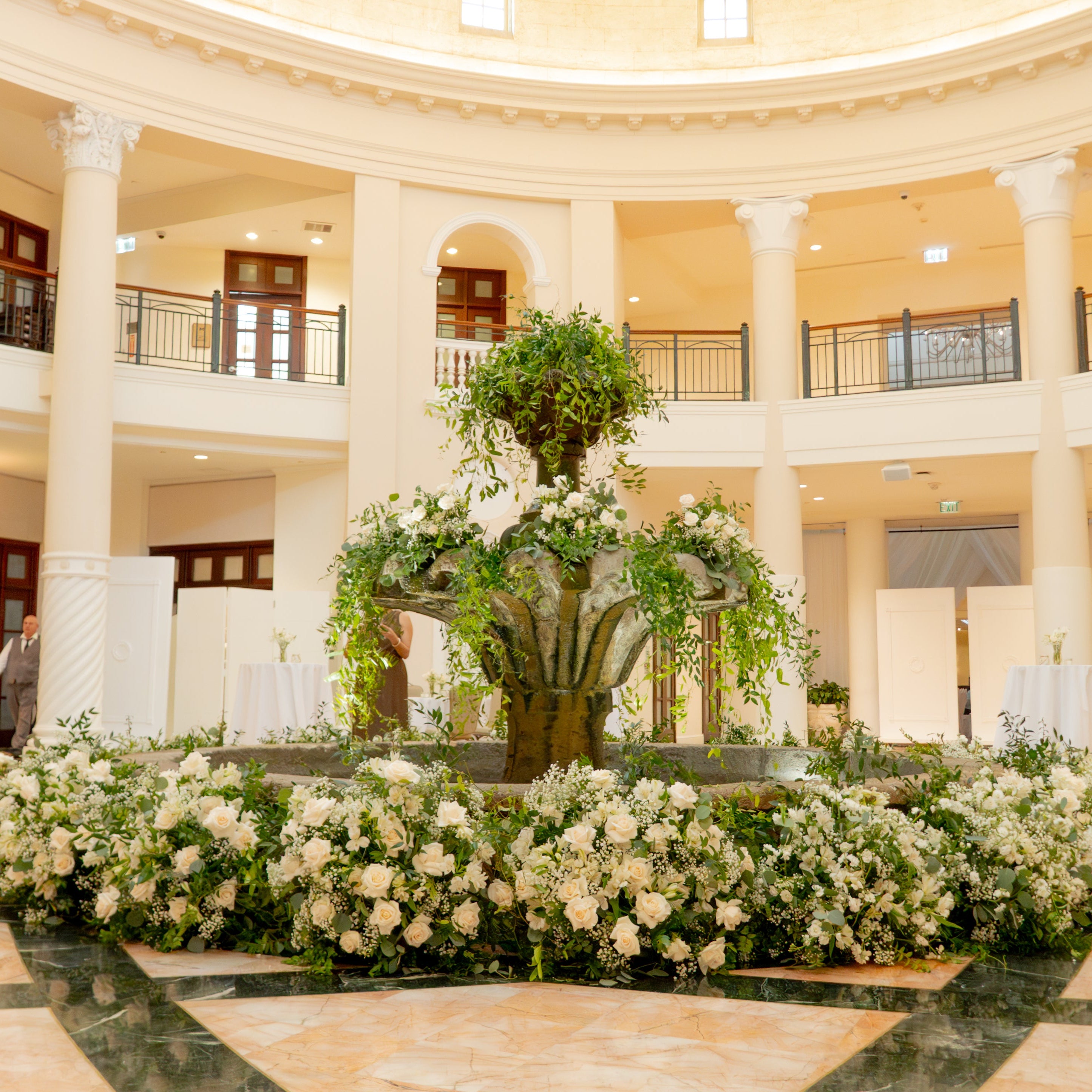 A large floral arrangement featuring a central green tree with white flowers and trailing foliage, set in a grand hotel lobby with white columns, white draped tables, and a marble floor.