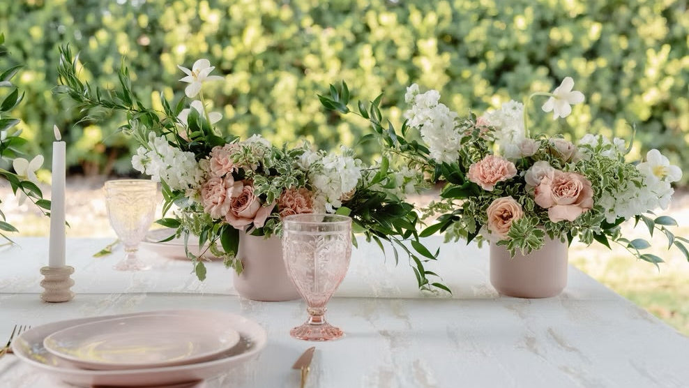 A styled outdoor table setting with two pink flower vases filled with white and blush pink flowers and green foliage, alongside pink glass goblets, a white candle, and plates on a white tablecloth, with a blurred green garden background.