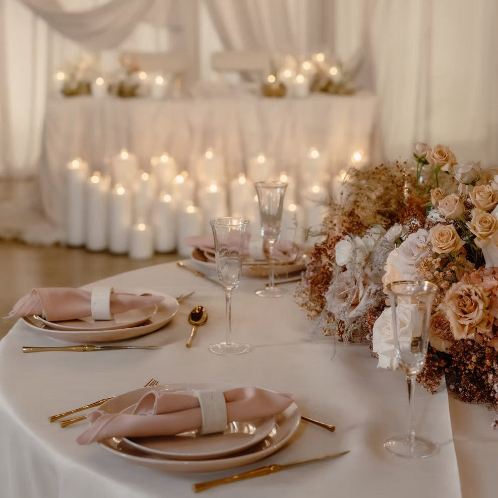 A beautifully styled wedding reception table setting with white tablecloths, blush pink napkins, gold flatware, clear glassware, and elegant floral centerpieces with dried flowers and roses under warm string lights.