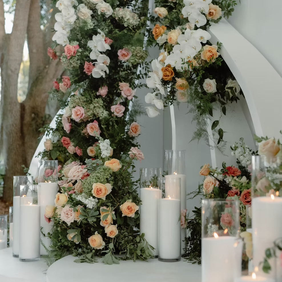 A white arched backdrop decorated with tall floral garlands in white, blush pink, peach, and orange, accented with green foliage and surrounded by white pillar candles in glass holders.