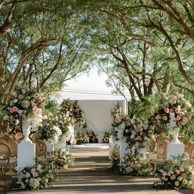 An outdoor wedding ceremony setup with a white tent backdrop, a central aisle, floral arrangements in pink, blush, and white, and rows of wooden chairs under tall trees.