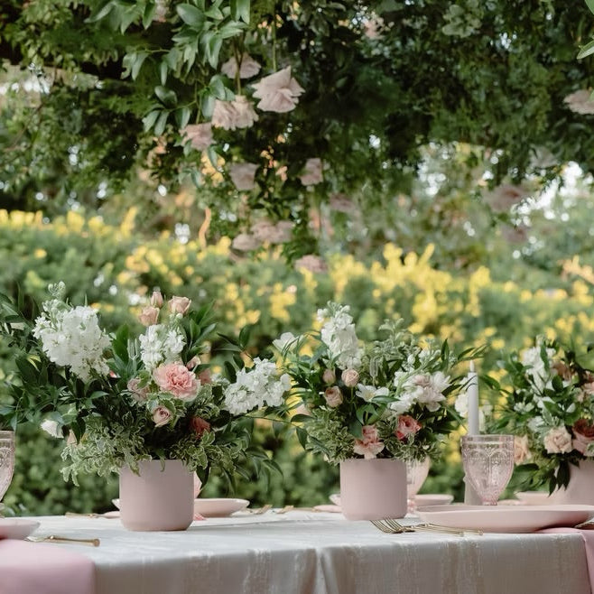 Outdoor wedding table setting with pink and white floral arrangements in pink pots, white plates, and clear glass candle holders under a tree canopy.