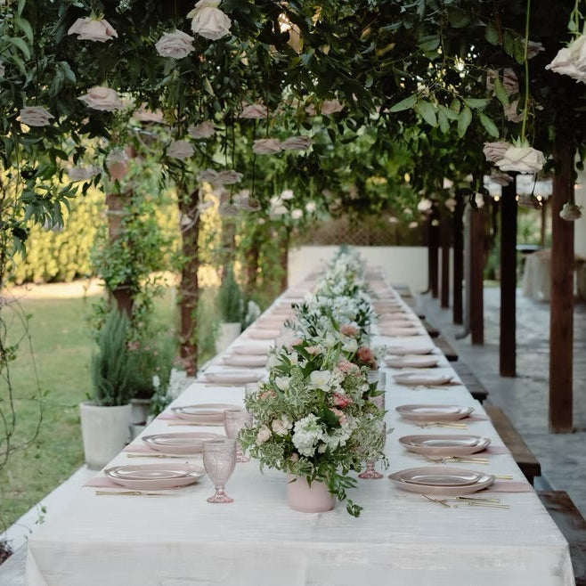 An outdoor wedding reception table setup under a canopy of flowering trees, featuring a long white table set with plates, clear glasses, and floral centerpieces.
