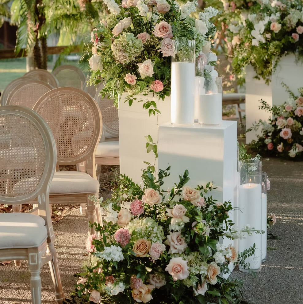 Outdoor wedding-style floral arrangements in soft pink, white, and green on white pedestals, with woven chairs and trees in the background.