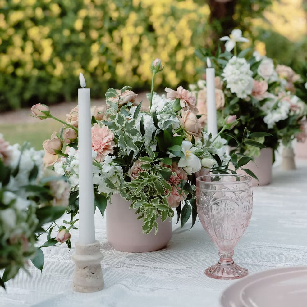 Outdoor garden table setting with pink flower arrangements in pale pink vases, white taper candles, and a pink textured glass, with a blurred green garden background.