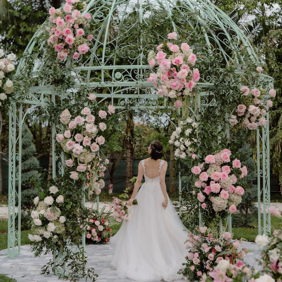 A bride in a white wedding gown stands under a mint green metal gazebo decorated with pink and white roses in an outdoor garden setting.