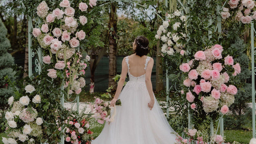 A bride in a white wedding gown stands under a mint green metal gazebo decorated with pink and white roses in an outdoor garden setting.