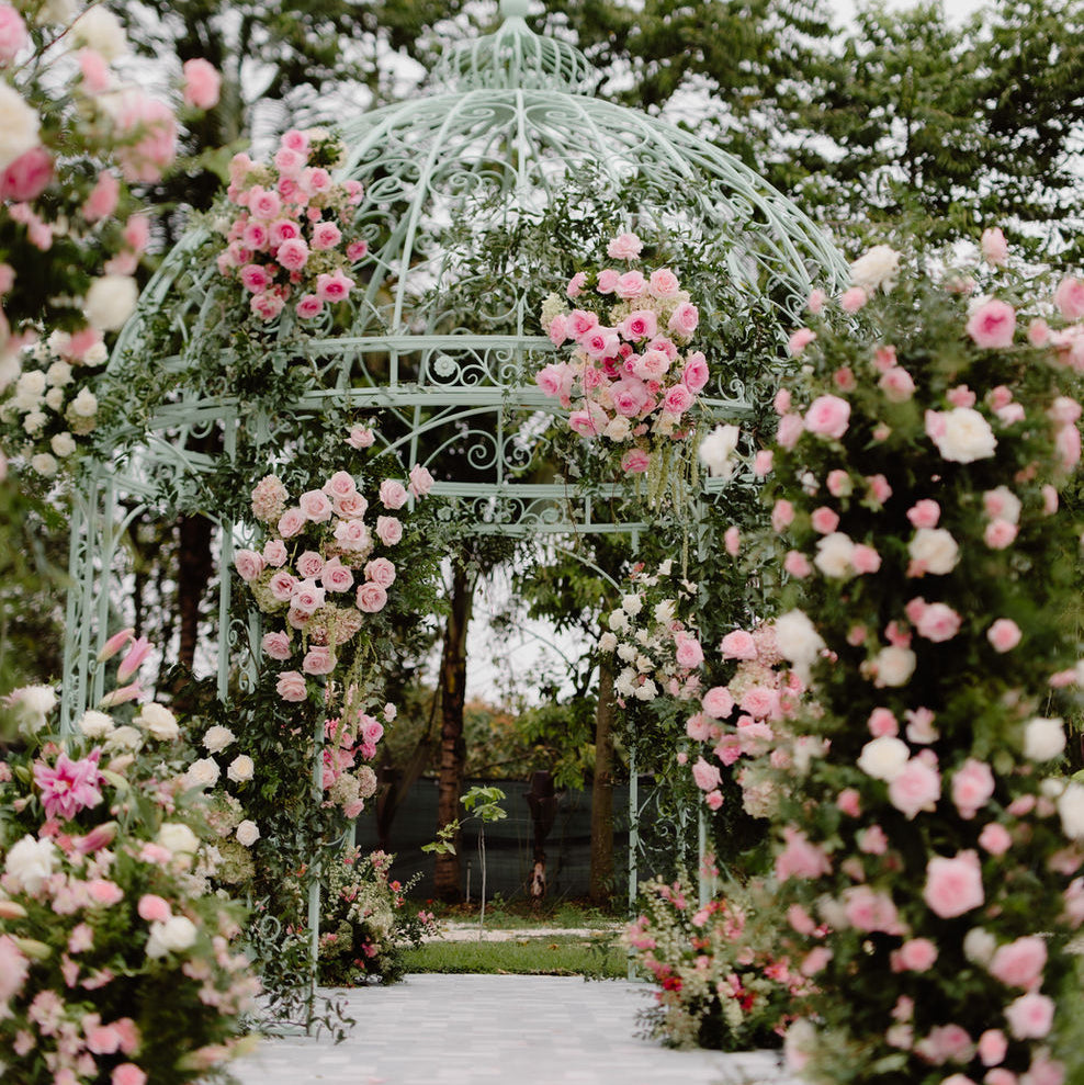 A decorative white metal gazebo archway surrounded by abundant pink and white roses, with a garden path leading through it and trees in the background.