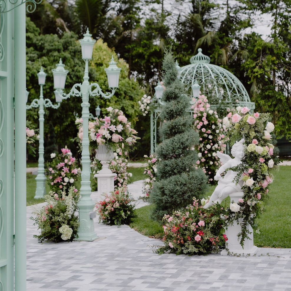 An outdoor garden wedding or event setup with a mint green decorative archway adorned with pink and white flowers, a matching dome structure, ornate white lampposts, and a stone pathway under a cloudy sky.