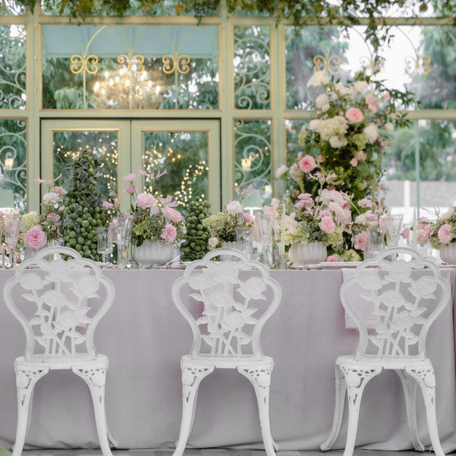 A beautifully decorated event table setup with a lavender tablecloth, three white ornate chairs, and multiple pink and white floral arrangements in front of a large windowed wall.