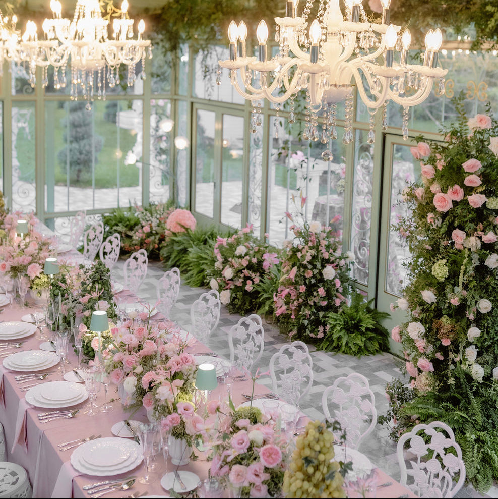 A beautifully decorated event space with long tables set in pink linens and white chairs, surrounded by abundant pink and white floral arrangements and greenery. Multiple white chandeliers hang from a high arched ceiling, with large windows in the background.