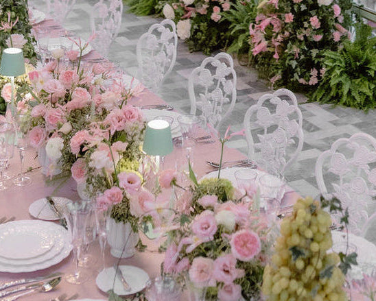 A beautifully decorated event space with long tables set in pink linens and white chairs, surrounded by abundant pink and white floral arrangements and greenery. Multiple white chandeliers hang from a high arched ceiling, with large windows in the background.