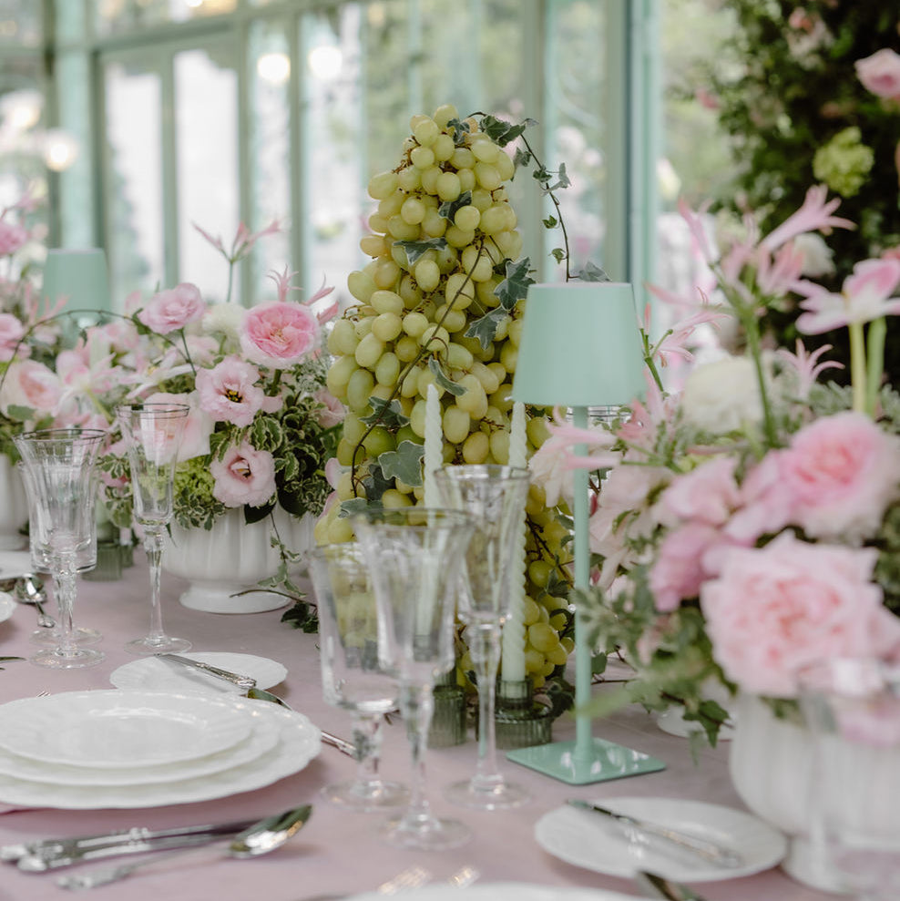 A styled dining table setup in a bright conservatory with pink floral arrangements, white plates, clear glassware, and a mint green table lamp. The scene is softly lit with large window panes in the background.