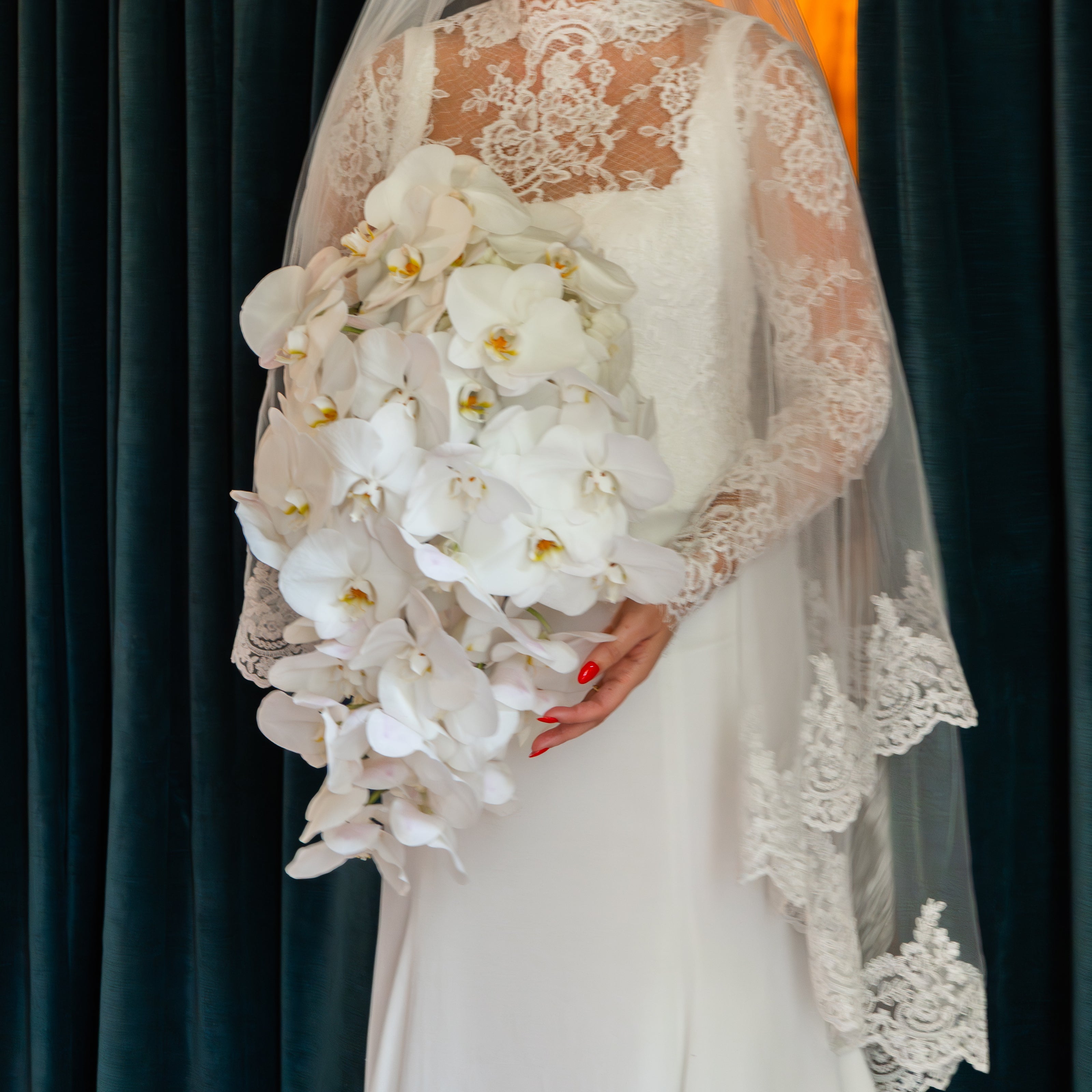 A bride in a white lace wedding dress and veil holding a large bouquet of white orchids, standing in front of dark teal curtains.
