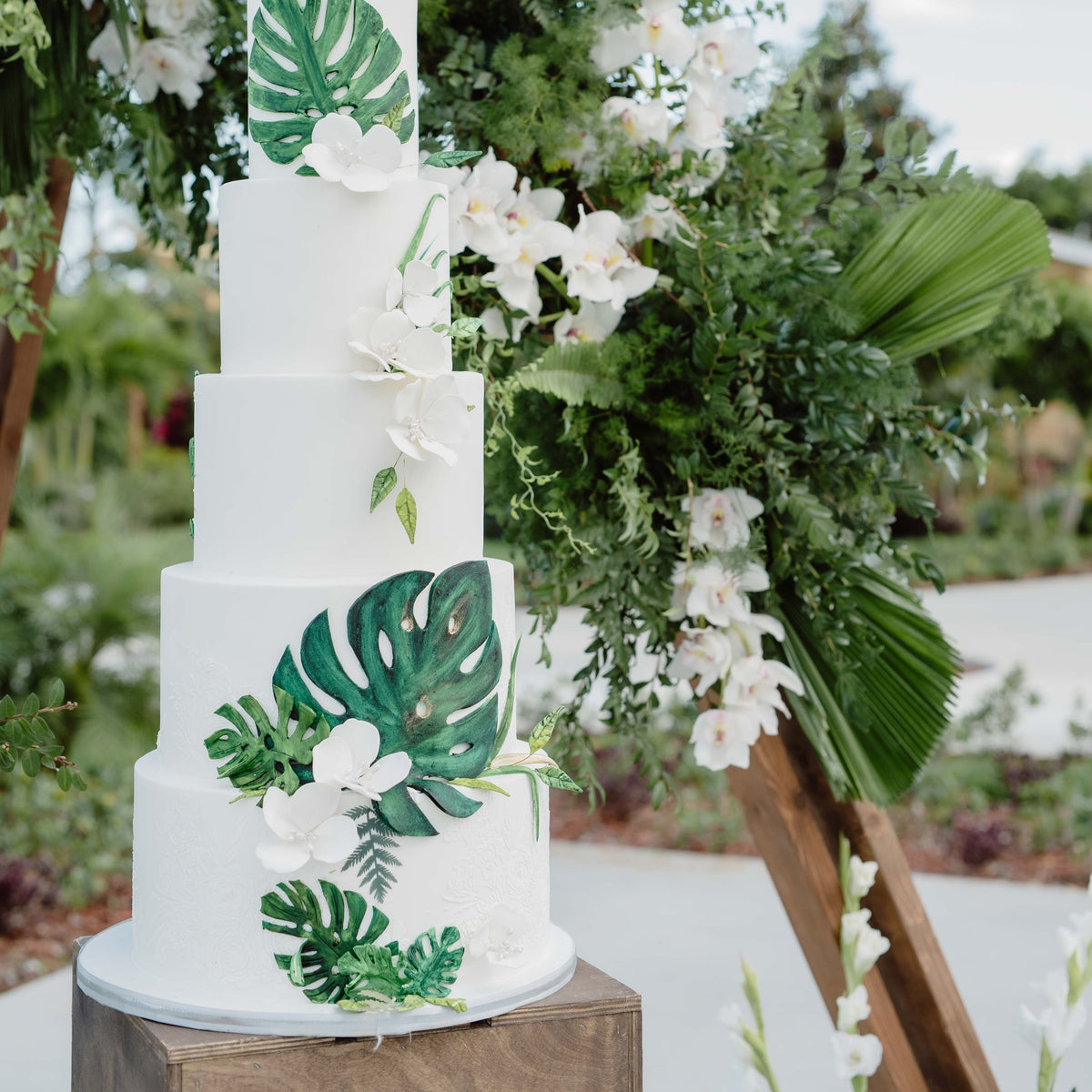 A four-tier white wedding cake decorated with green tropical monstera leaves and white orchids, displayed outdoors next to a wooden arch with lush greenery and white flowers.