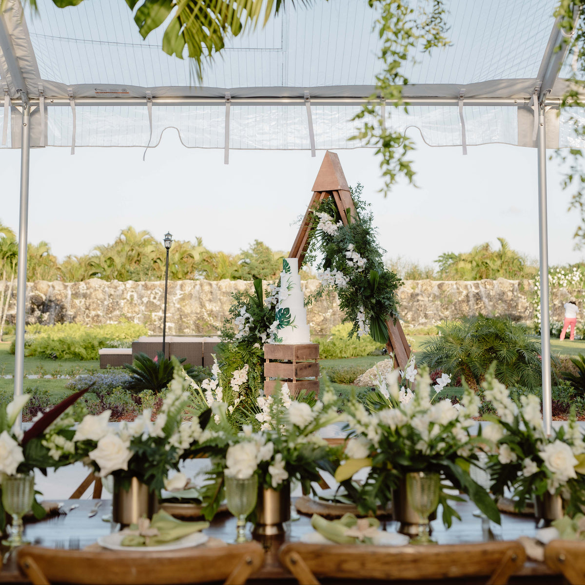 Outdoor wedding reception table setup under a clear tent canopy, decorated with lush green tropical foliage hanging overhead, white floral centerpieces, and a wooden arch in the background.