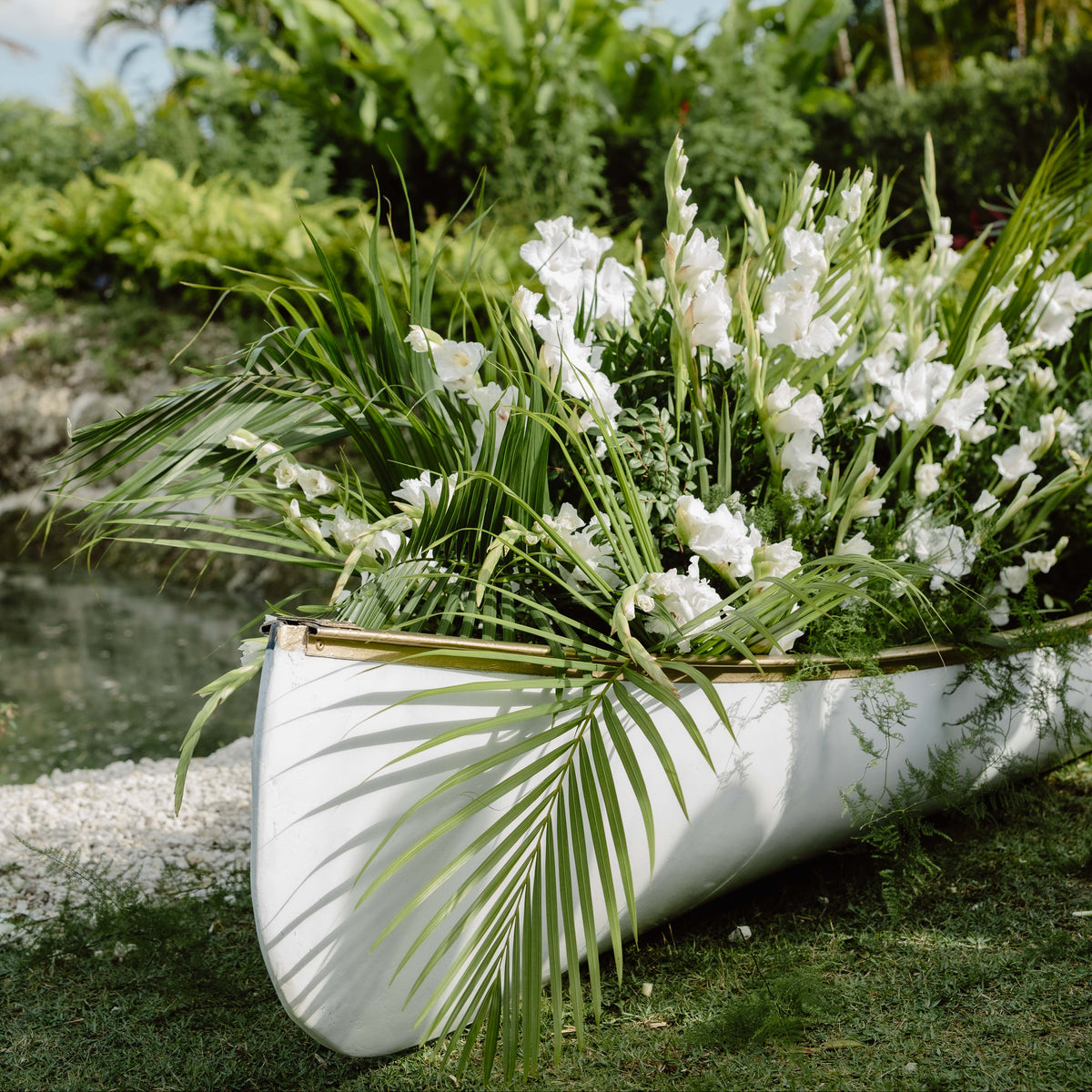 A white canoe filled with lush green tropical foliage and white flowers, resting outdoors on grass near palm trees and water under a blue sky.