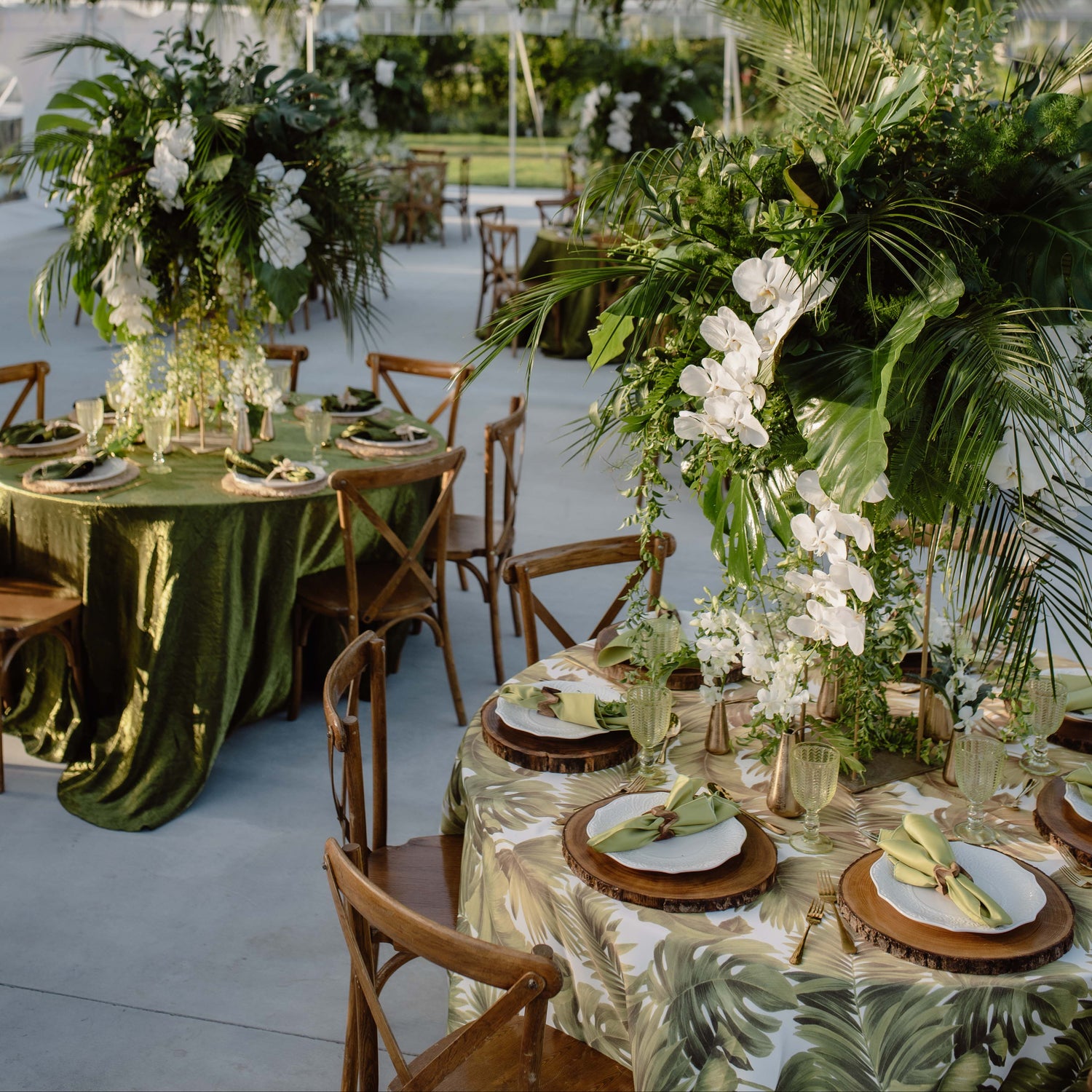 An elegant event setup inside a glass marquee with lush green tropical floral arrangements, hanging wooden lanterns, and tables set with white flowers, green linens, and wooden chairs.