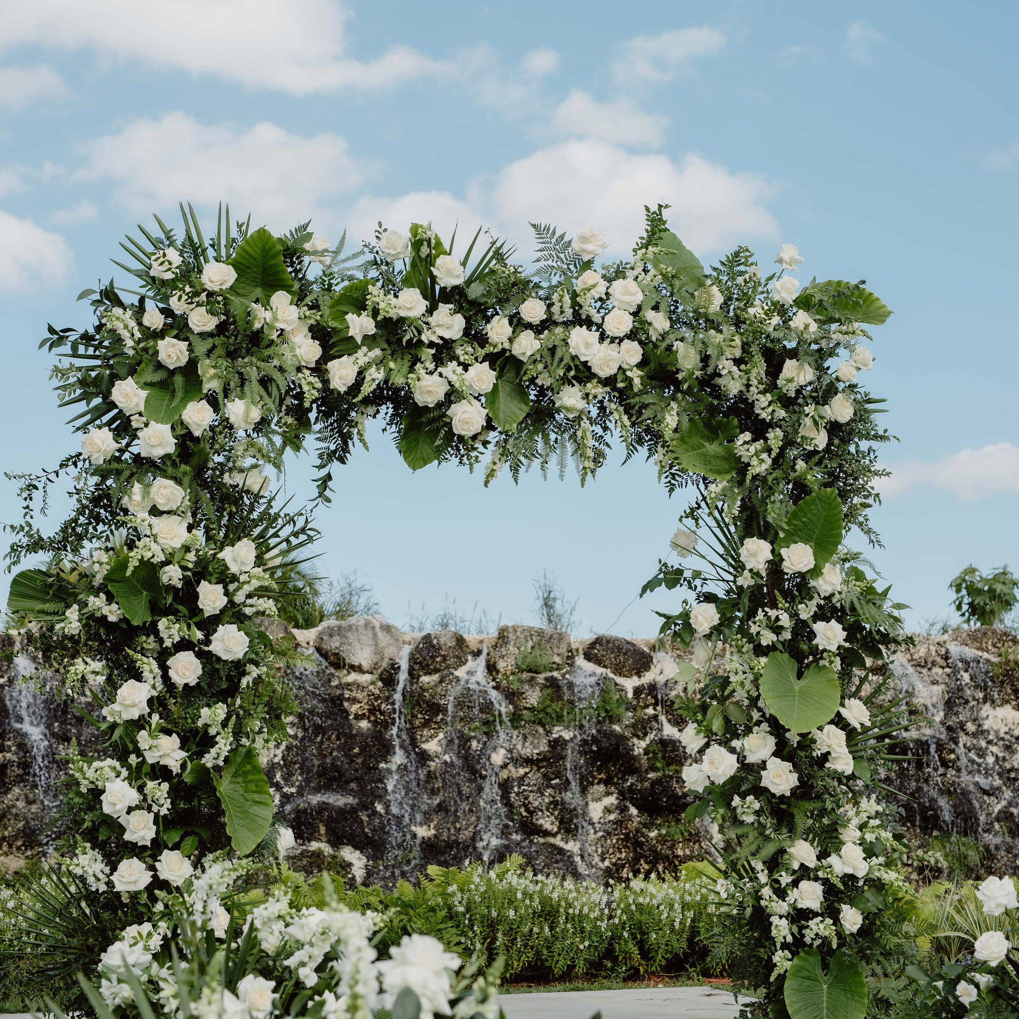 A large floral wedding arch decorated with white flowers and green foliage, set outdoors in front of a waterfall and blue sky.