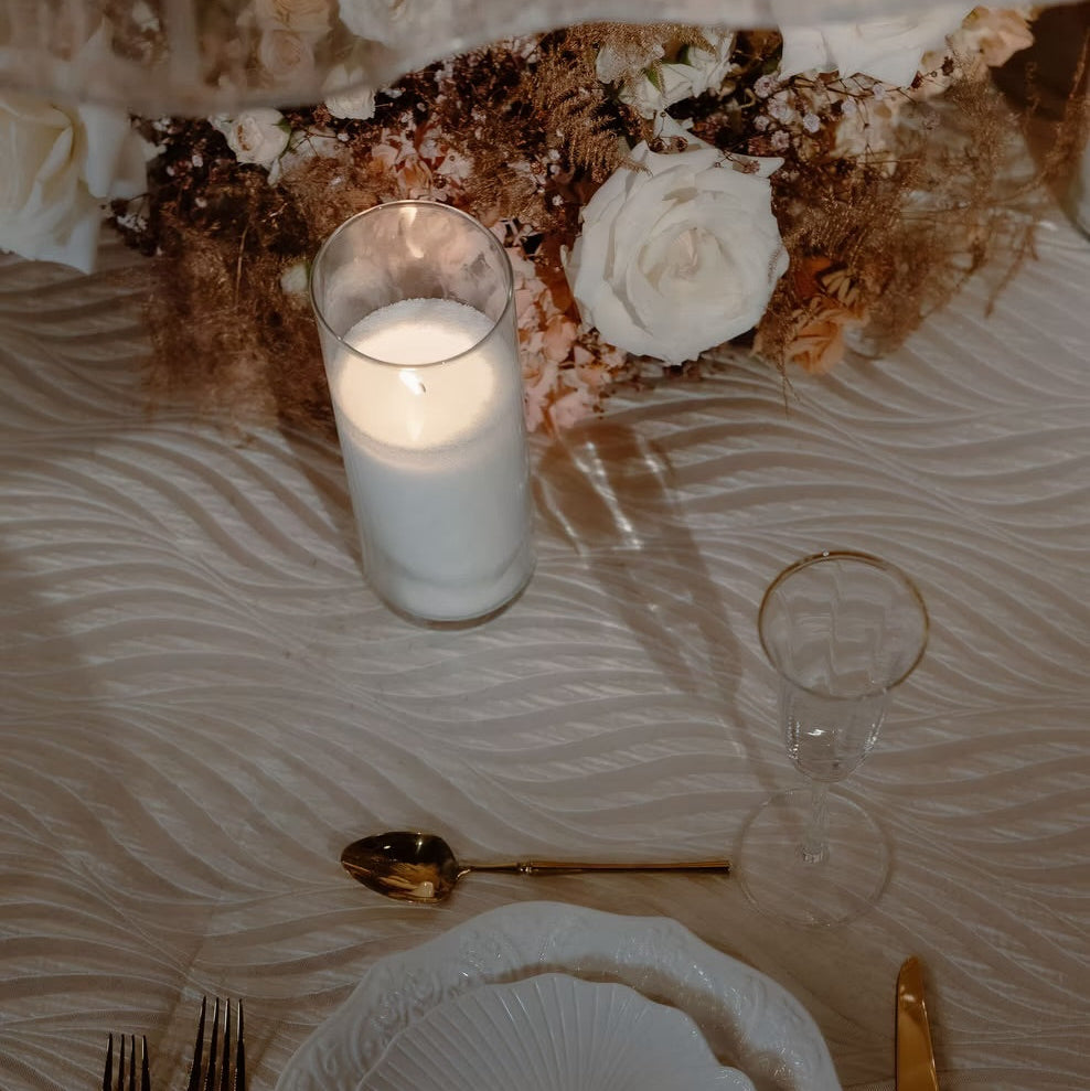 A styled wedding table setting featuring a white textured charger plate and matching bowl, gold flatware, a lit white candle in a glass holder, and a clear wine glass on a beige patterned tablecloth.