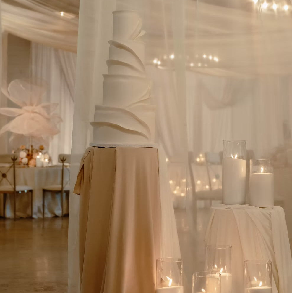 A tall, tiered white wedding cake displayed on a draped beige fabric pedestal in an indoor event space with sheer white drapery, lit candles in glass holders, and tables in the background.