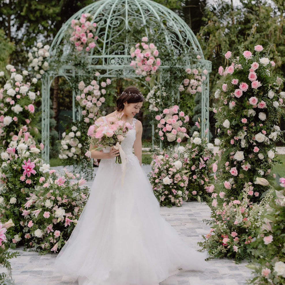 A bride in a white wedding gown stands under a mint green metal gazebo decorated with pink and white roses, holding a bouquet in a garden setting.