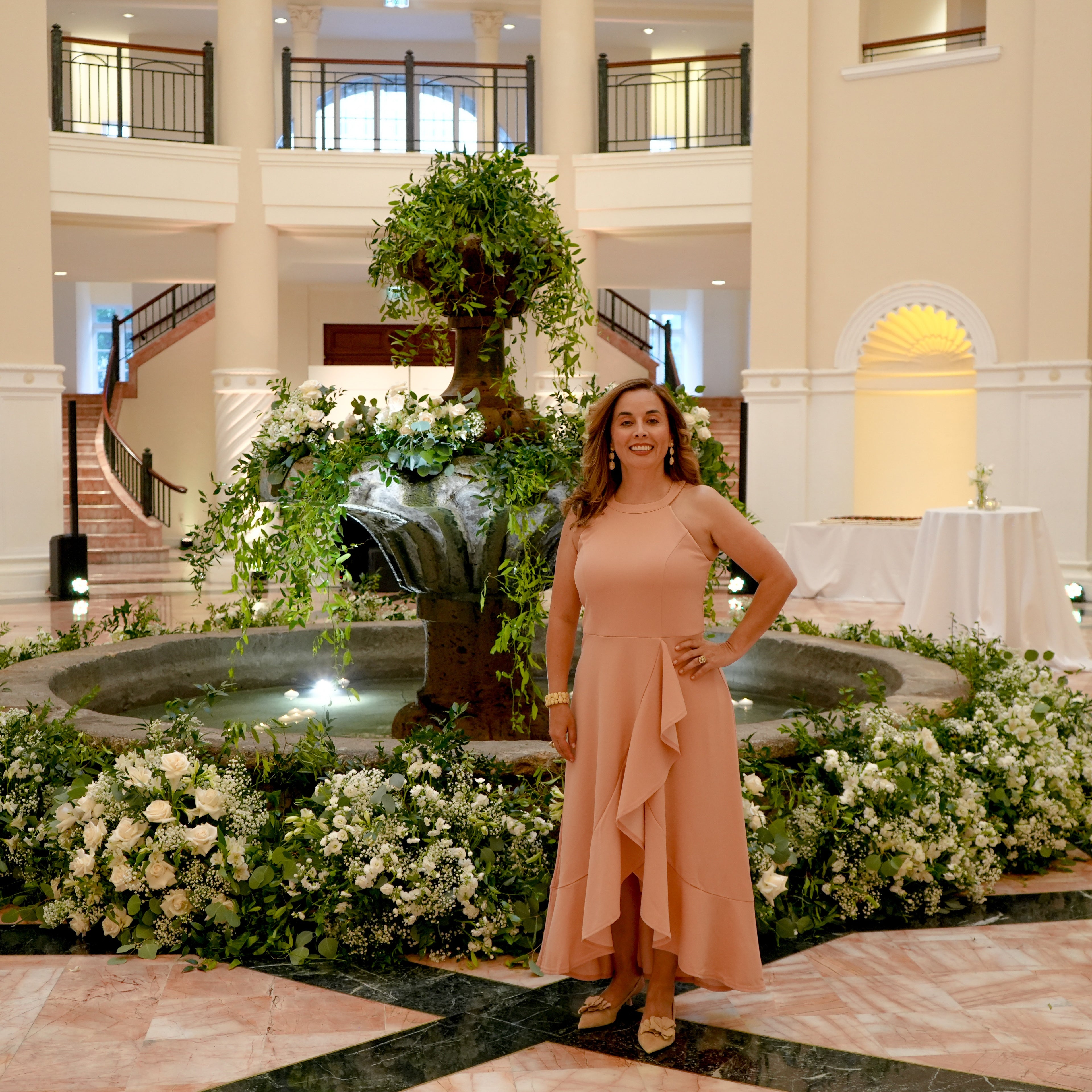 A woman stands in a grand hotel lobby in front of a central fountain and floral arrangements, wearing a peach ruffled midi dress with a high-low hem and sleeveless design.