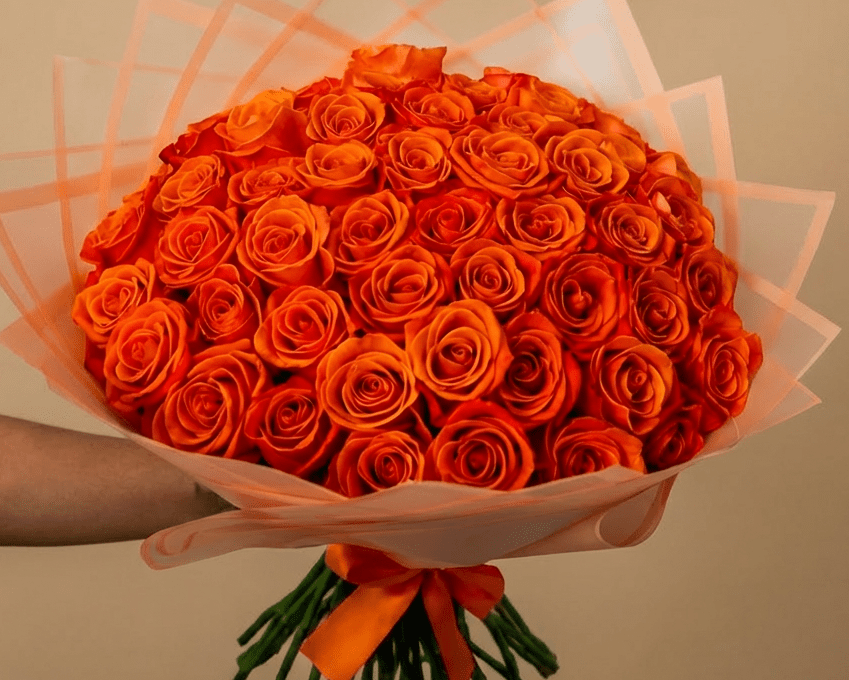A large bouquet of vibrant orange roses wrapped in pale pink tissue paper and tied with an orange ribbon.