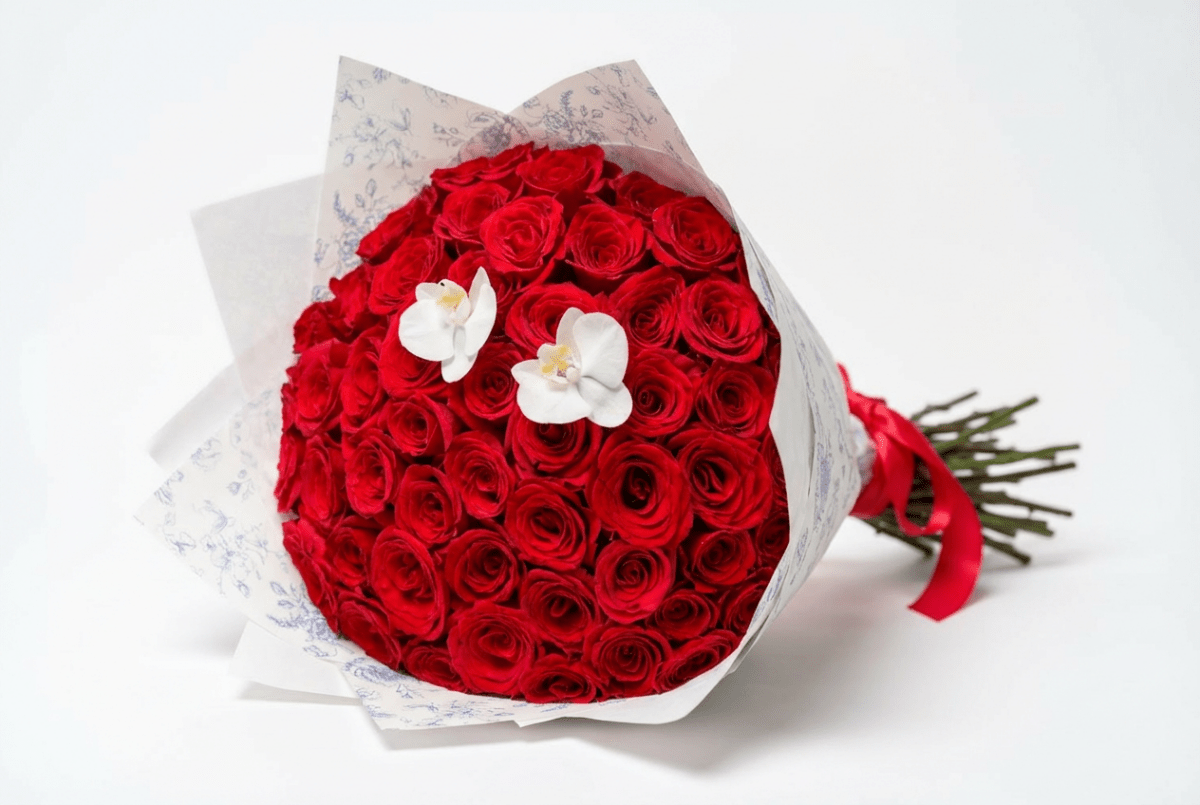 A round bouquet of red roses wrapped in white patterned paper with two white orchid flowers on top and a red ribbon.