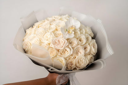 A large round bouquet of white and cream roses wrapped in white tissue paper, with a single white orchid flower on top, held by a hand against a plain light gray background.