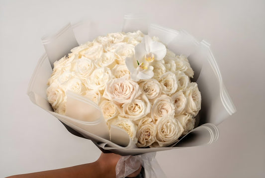 A large round bouquet of white and cream roses wrapped in black paper, with a single white orchid flower on top.