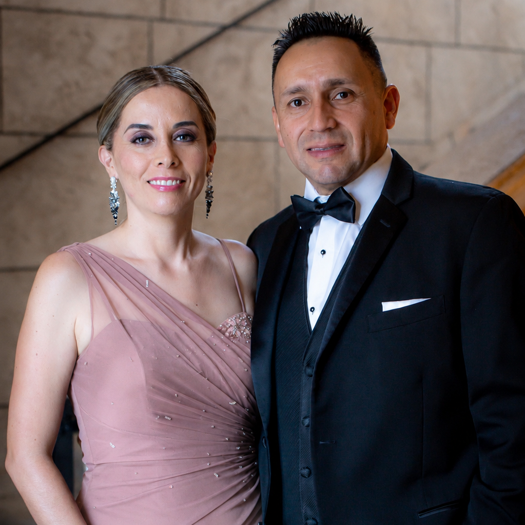 A man and a woman posing in formal evening wear. The woman wears a sleeveless, draped dusty rose pink gown with subtle sparkles, and the man wears a classic black tuxedo with a white shirt, black bow tie, and white pocket square.
