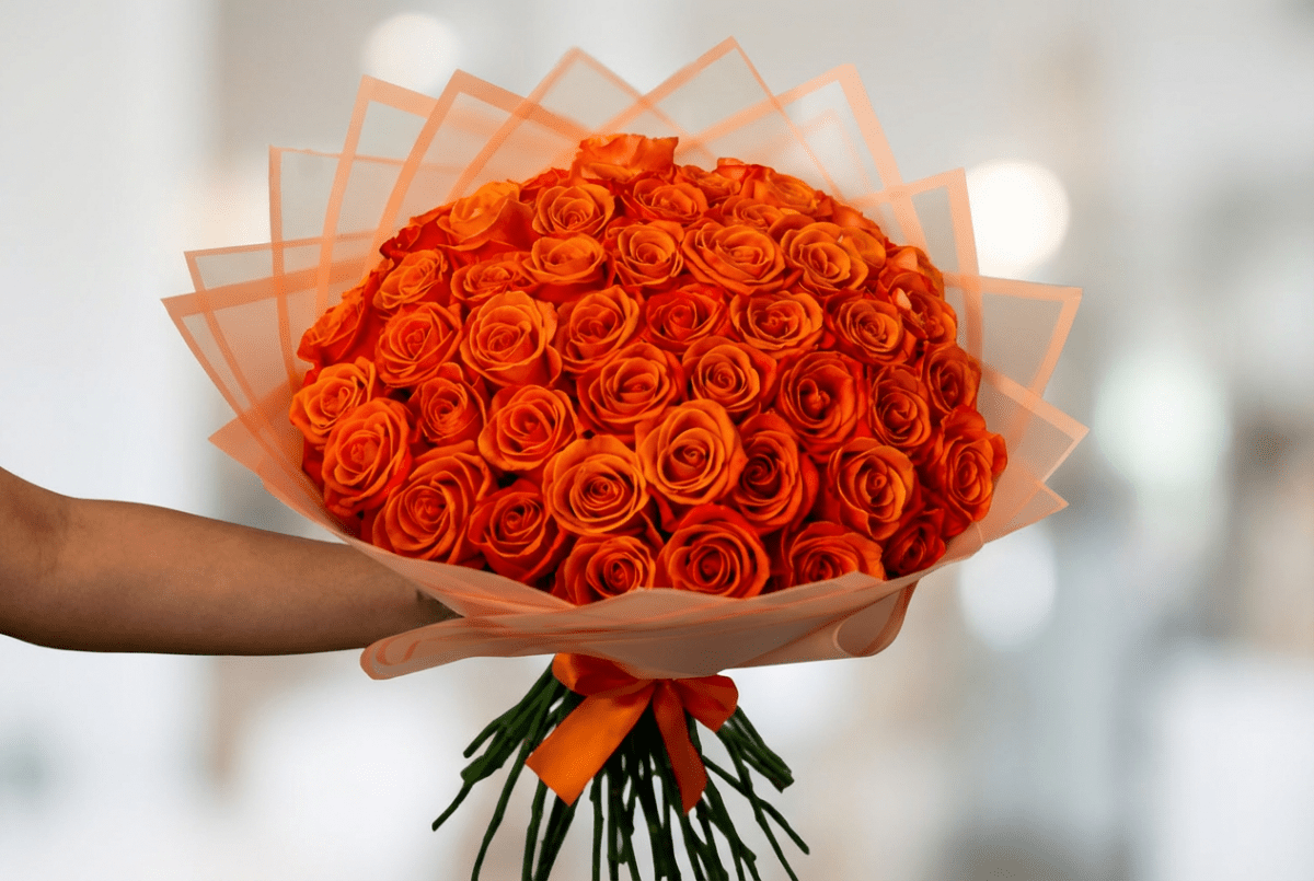 A large bouquet of vibrant orange roses wrapped in peach-colored tissue paper and tied with an orange ribbon, held by a person's arm against a soft, blurred white background.