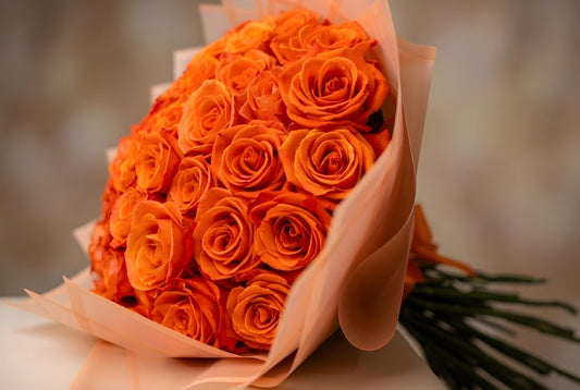 A close-up of a bouquet of vibrant orange roses wrapped in light peach paper, against a soft, blurred neutral background.
