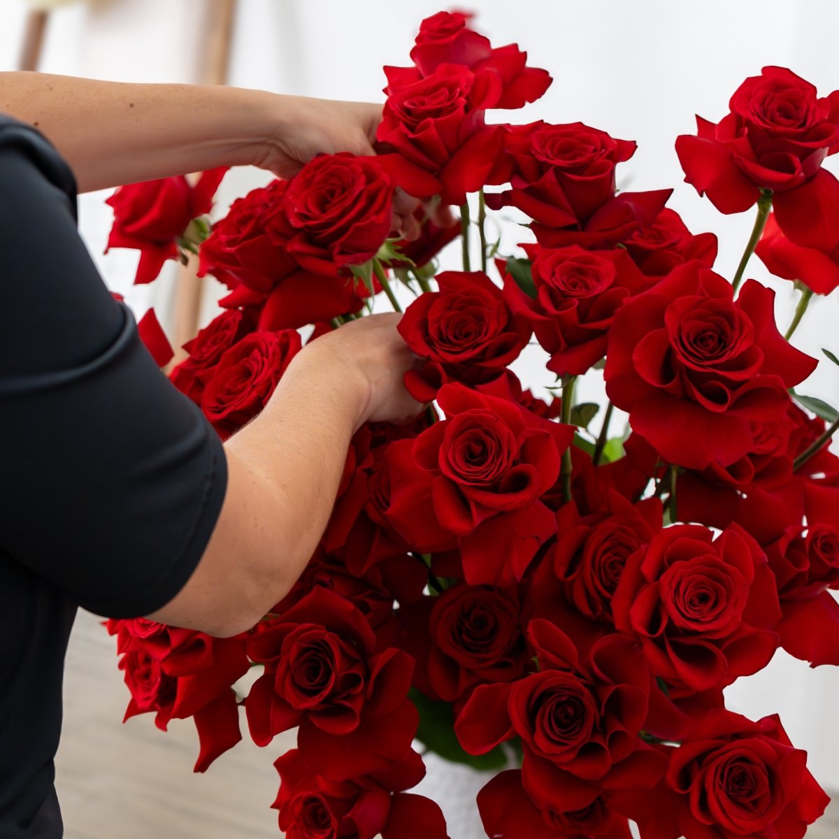 A close-up of a person's hands arranging a large bouquet of vibrant red roses, with their black short-sleeve shirt visible.