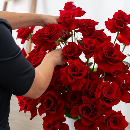 A close-up of a person's hands arranging a large bouquet of vibrant red roses, with their black short-sleeve shirt visible.