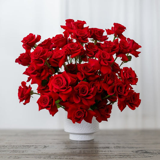 A dense bouquet of vibrant red roses arranged in a small white ceramic pot, set on a rustic wooden tabletop.