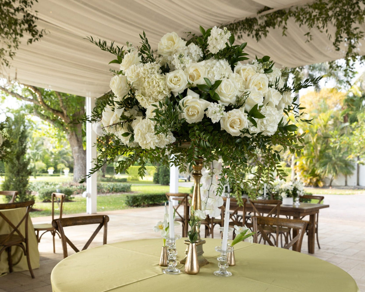 A round table covered with a pale yellow-green tablecloth, set under a draped white tent with wooden chairs and a large white rose floral centerpiece.