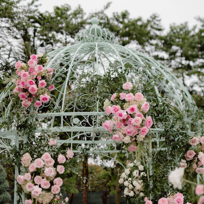 A pastel mint green metal gazebo decorated with abundant pink and white roses and green foliage, set outdoors against a soft-focus background of trees.