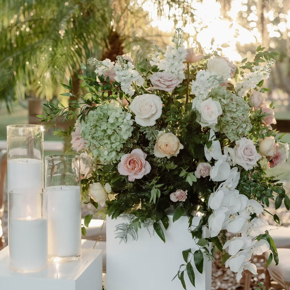 Floral arrangement with white and pink flowers in a vase, surrounded by candles on a pedestal outdoors.