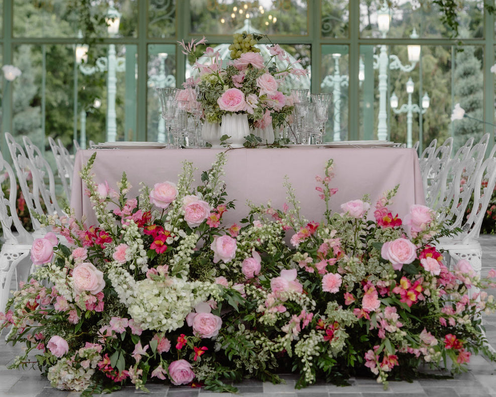 A beautifully decorated event space with a long table covered in a pale pink cloth, surrounded by abundant floral arrangements in pink, red, and white with green foliage. Multiple ornate white chandeliers hang from a green canopy overhead, with white chairs arranged around the table in a large glass conservatory.