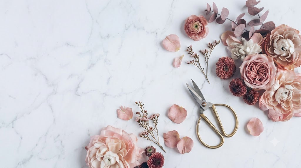 A styled flat-lay of dried and fresh pink, peach, and burgundy flowers with green eucalyptus stems, gold-handled scissors, and scattered petals on a white marble surface.