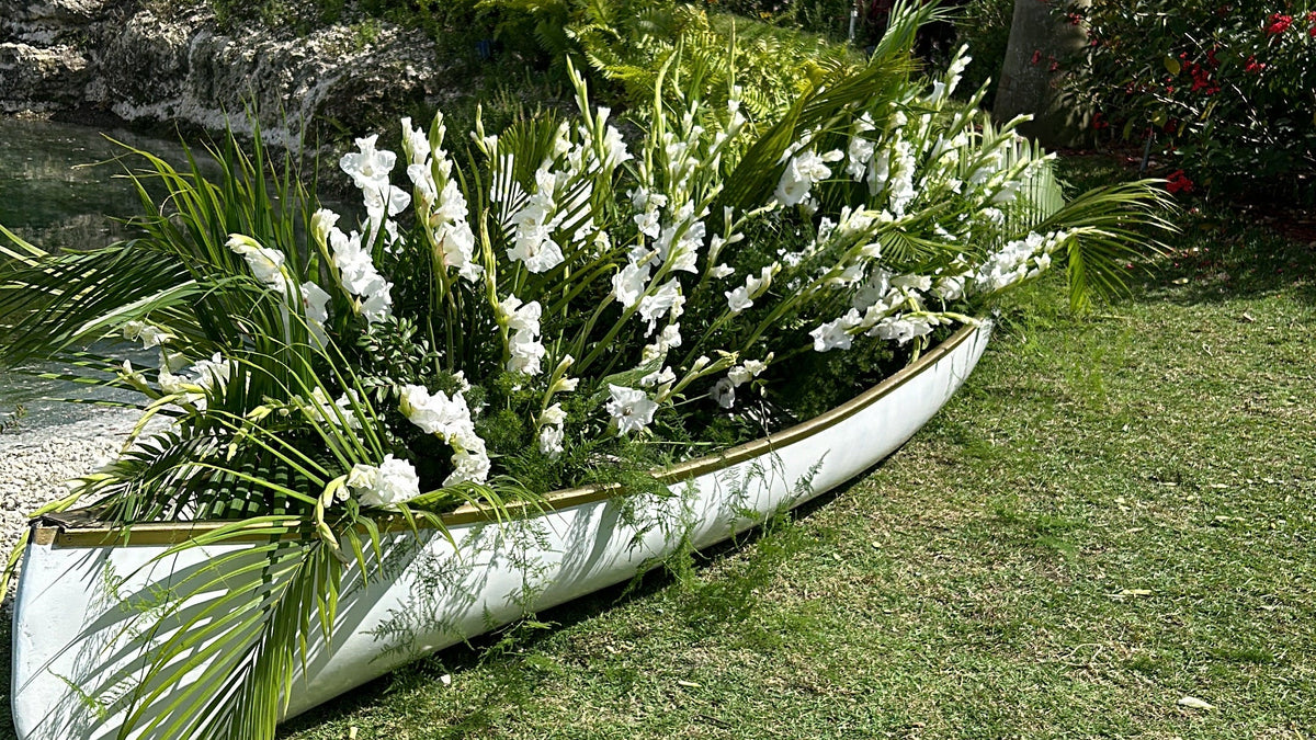 A white decorative boat-shaped planter filled with white flowers and green palm fronds, placed on a green lawn outdoors with tropical plants and a blue sky in the background.