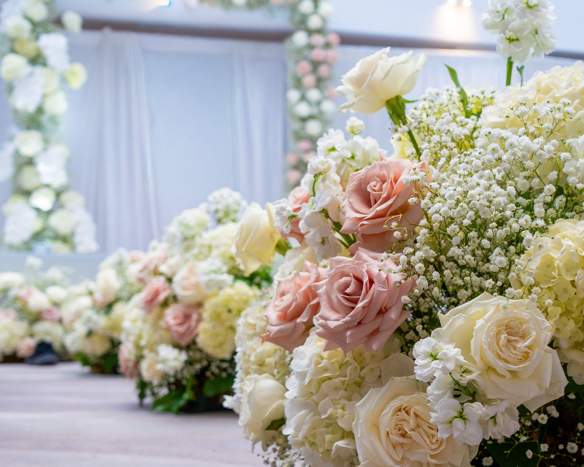 Large floral arrangements of blush pink roses, cream roses, baby's breath, and white hydrangeas placed along an aisle in an indoor event space with draped white fabric and floral arches in the background.