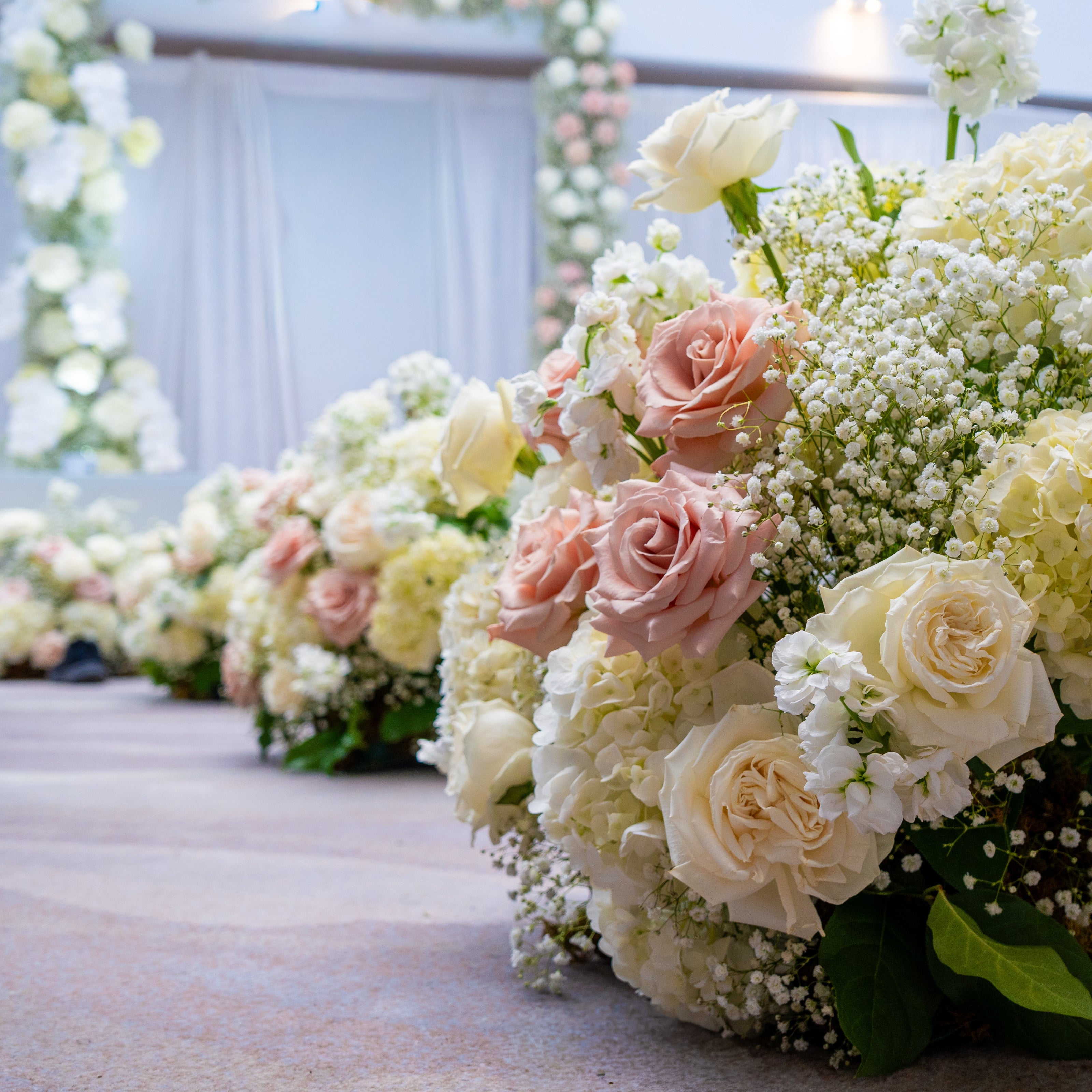 Large floral arrangements of blush pink roses, cream roses, baby's breath, and white hydrangeas placed along an aisle in an indoor event space with draped white fabric and floral arches in the background.