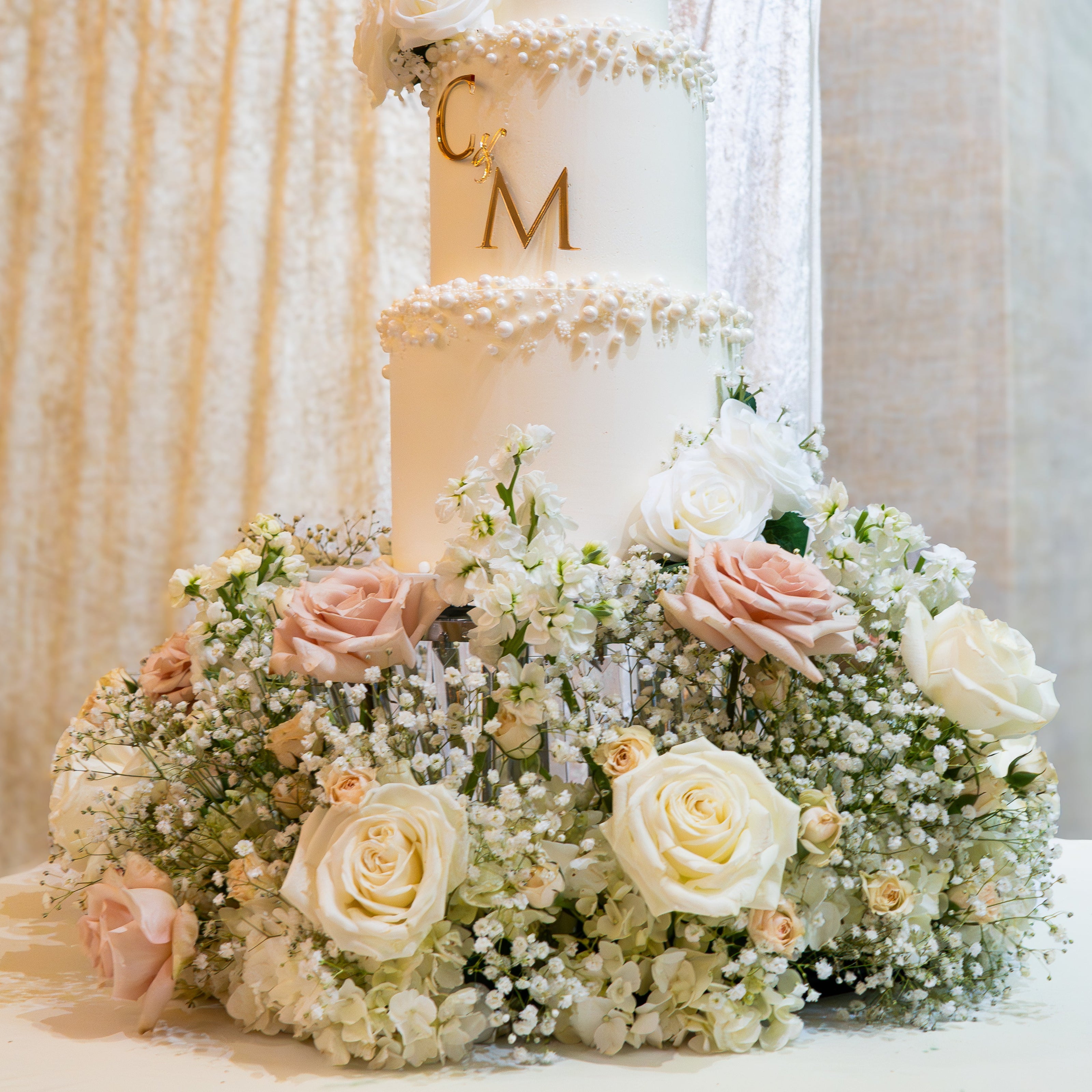 Three-tiered wedding cake with floral decorations and 'CM' initials against a beige curtain background.